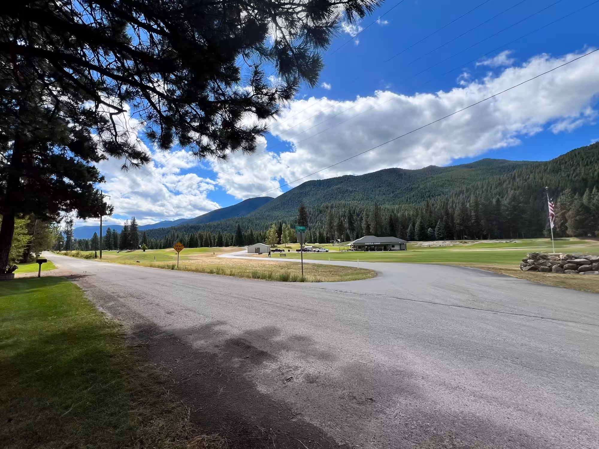 A quiet country road runs past broad mown golf turf, where a long low building with a dark hipped roof and a deep covered porch stands across the way with a few pickups parked on the grass nearby and an American flag on a tall pole off to the right. Dense evergreen forest and timbered mountains rise behind it under a bright blue sky stacked with white clouds.
