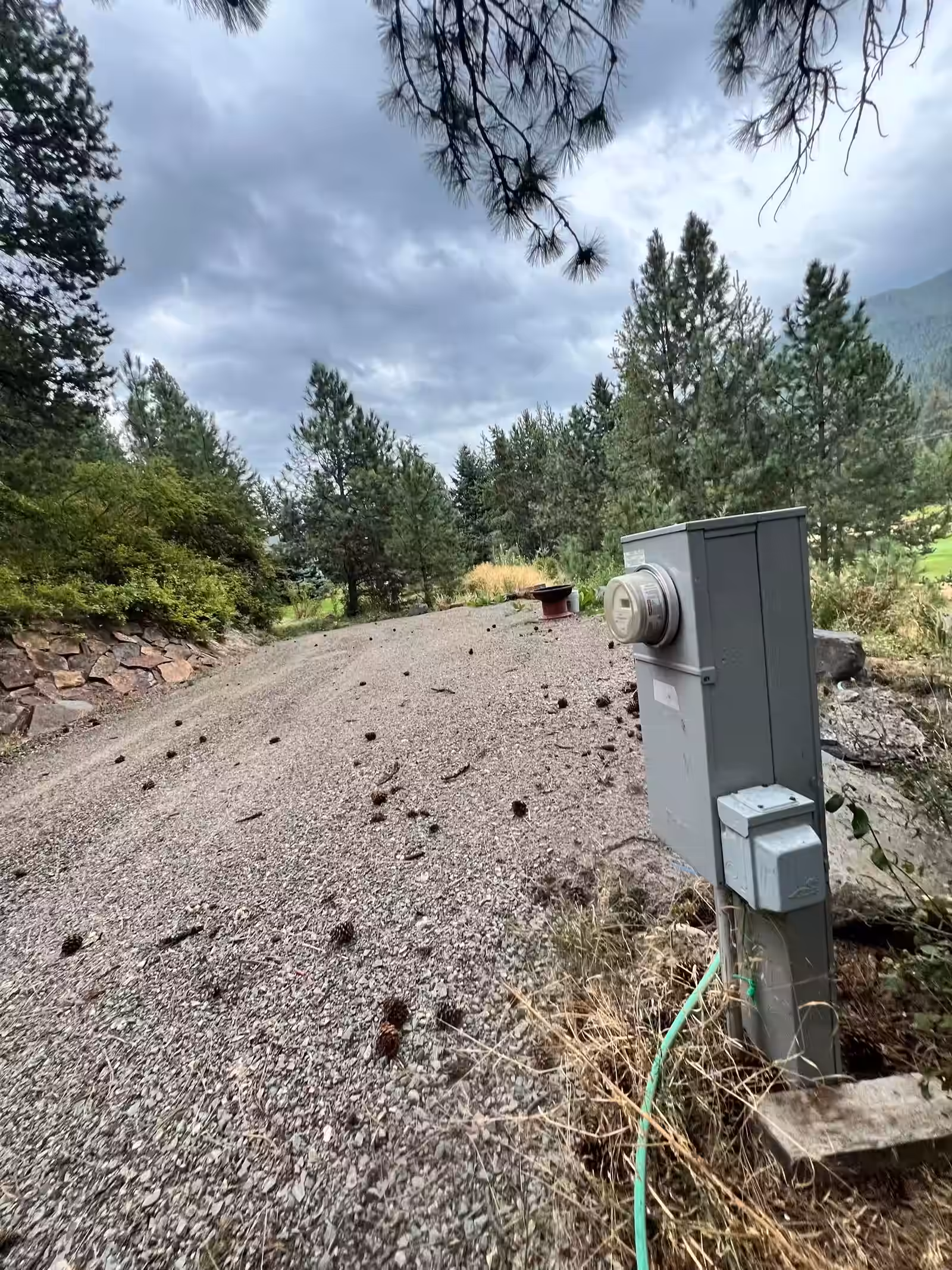 A grey power pedestal with weatherproof outlet covers stands at the edge of a gravel pad, a green hose running down from its base and a low capped pipe riser standing at the pad's edge beyond it. Pine cones dot the gravel, a dry-stacked stone wall runs along one side, and tall pines and a forested slope rise beyond under an overcast sky.