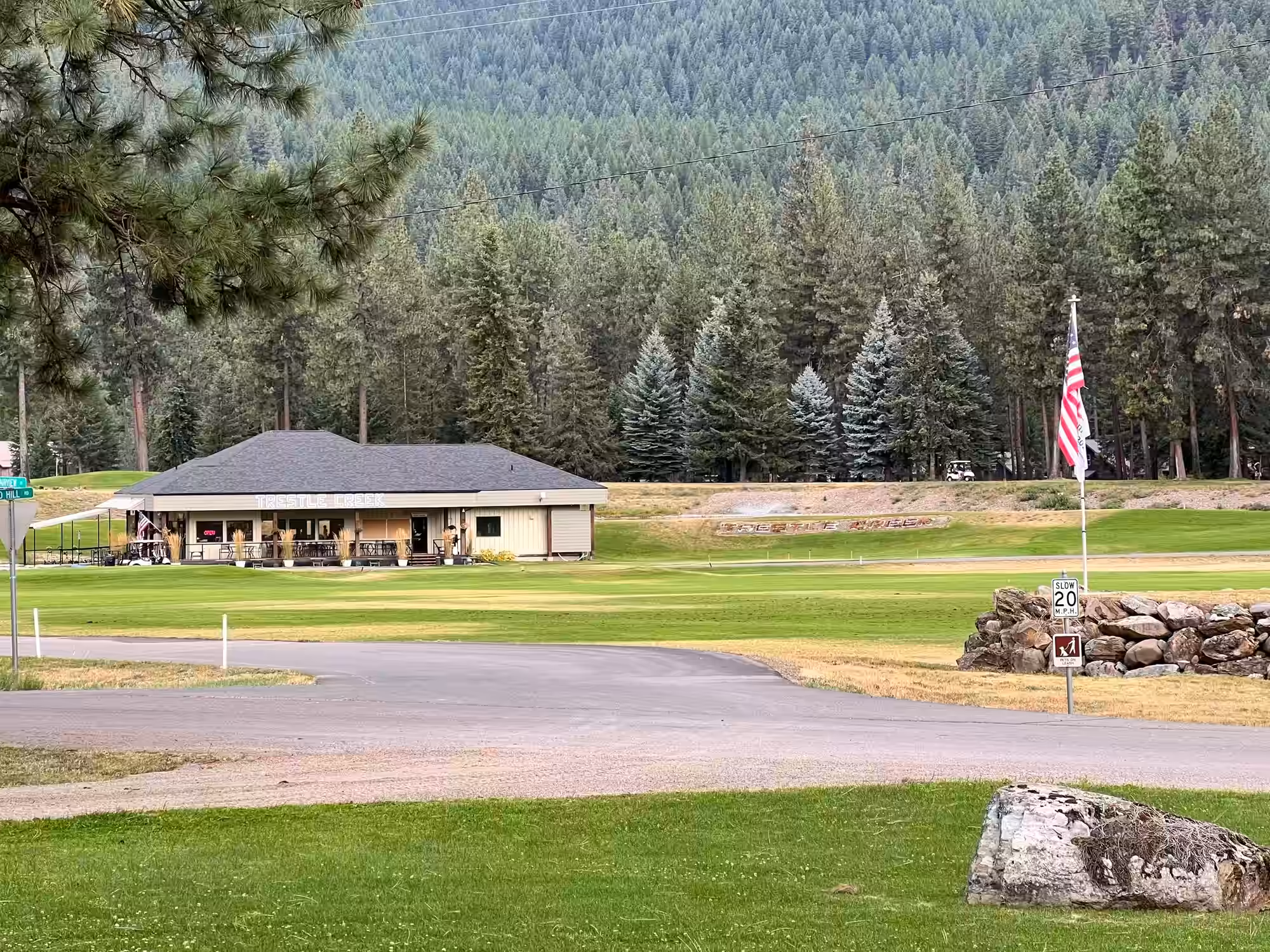 The Trestle Creek golf clubhouse sits just across the road, a long cream-sided building under a broad dark hipped roof, its covered porch lined with small tables, chairs and tall planters of ornamental grass. Close-mown green turf, a rock-edged bank and a flagpole flying the Stars and Stripes spread out in front of it, with a steep wall of evergreen forest rising behind.
